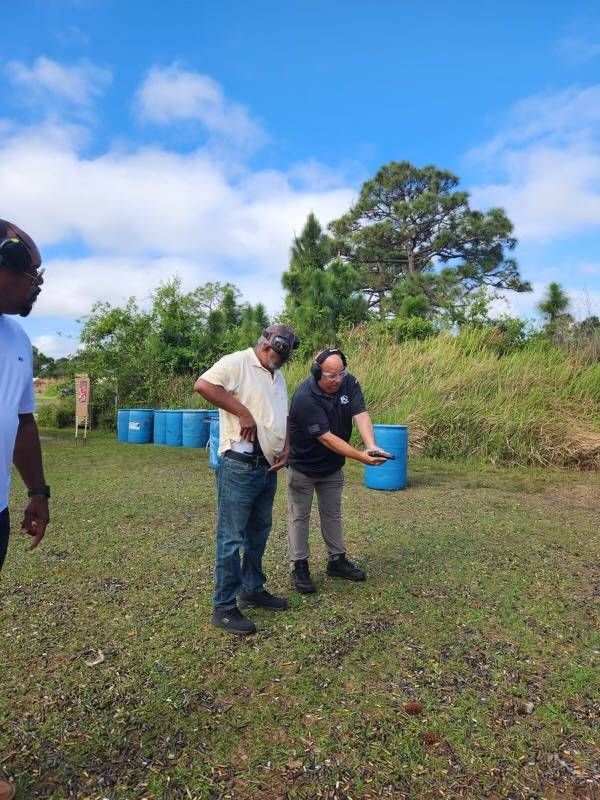 A man in jeans and a beige shirt is being instructed by a person in a black shirt at an outdoor shooting range.