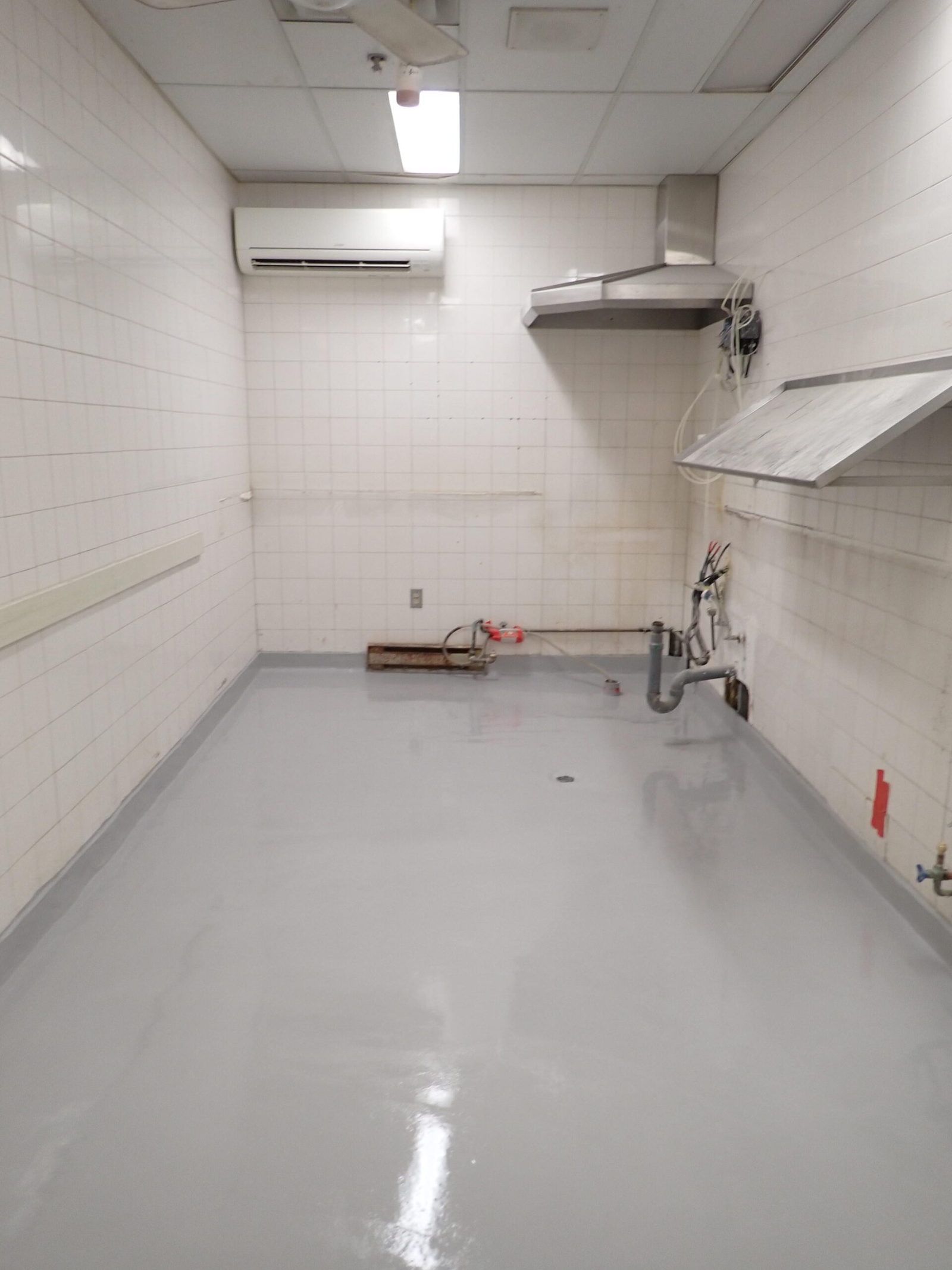 Cuisine vide avec sol gris, murs carrelés blancs et hotte aspirante.