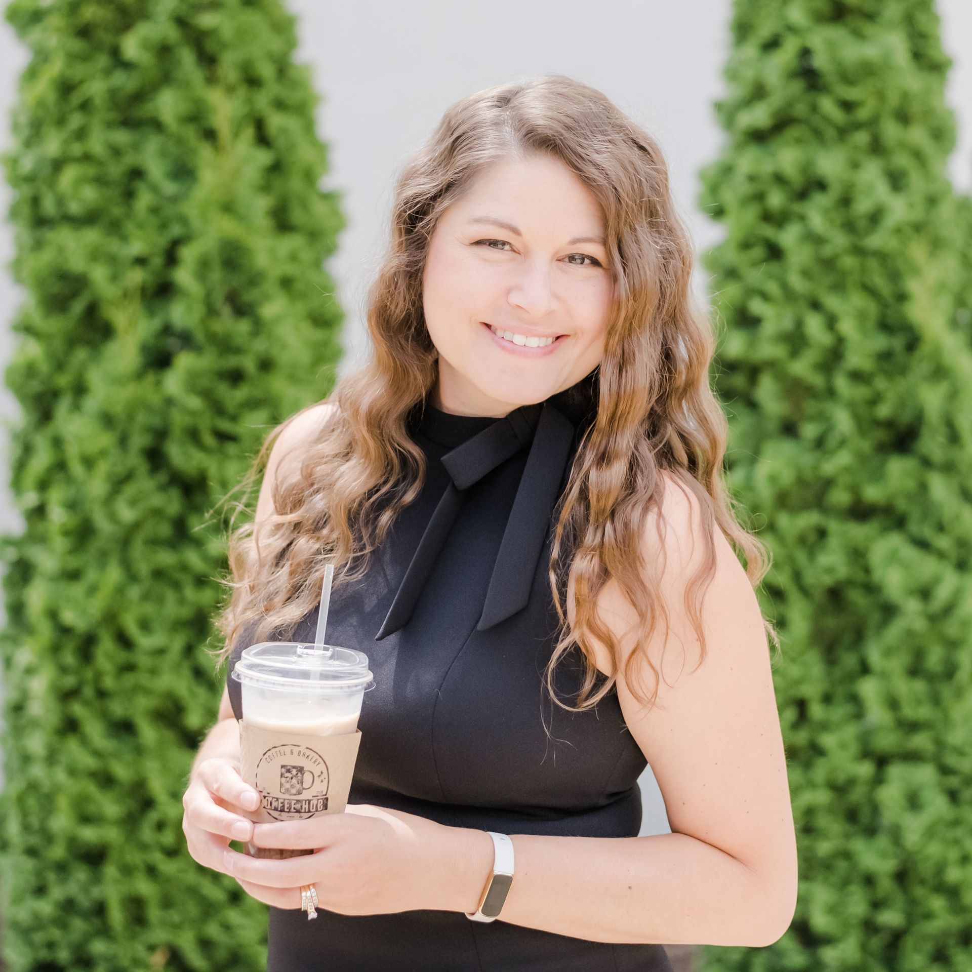 Woman with wavy brown hair smiles, holding iced coffee. She wears a black top, standing outside near tall green bushes.