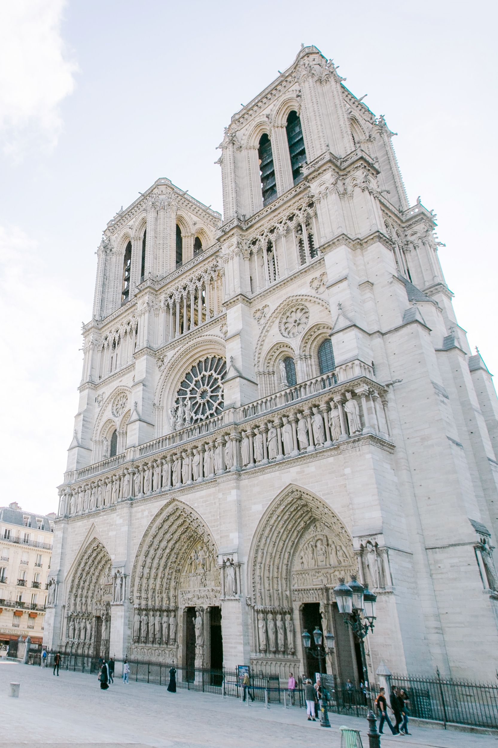 Notre Dame Cathedral in Paris, France. White stone facade, Gothic architecture. Cloudy sky. People walking nearby.