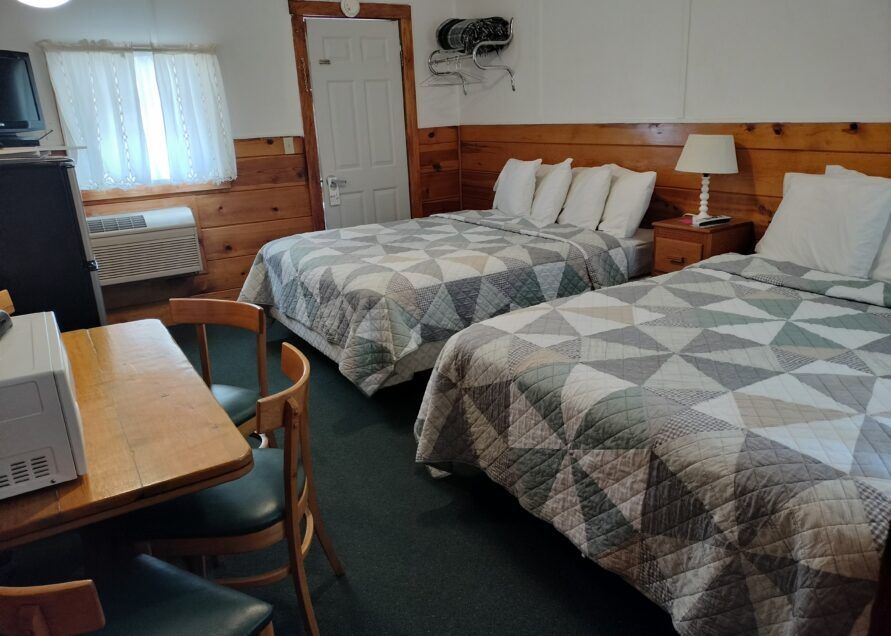 A small motel room with two double beds, a desk, and a microwave. Wood-paneled walls and green carpet.