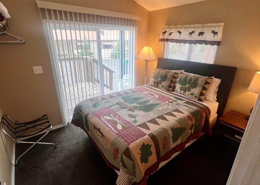 A bedroom with a bed featuring a quilt with trees and animals, a sliding door to a deck, and a small side chair.