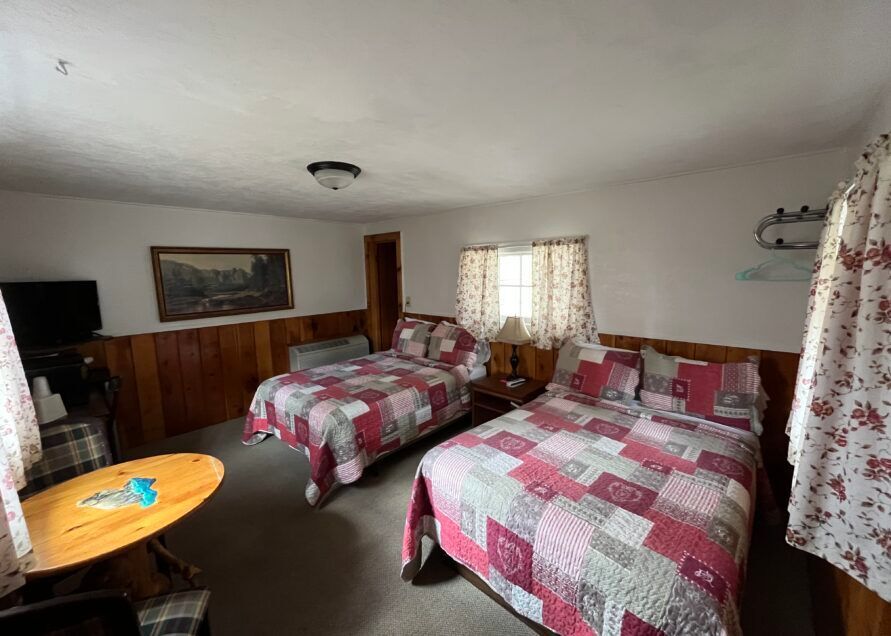 A motel room with two double beds, a small table, and a TV. The beds have red and gray patterned quilts.