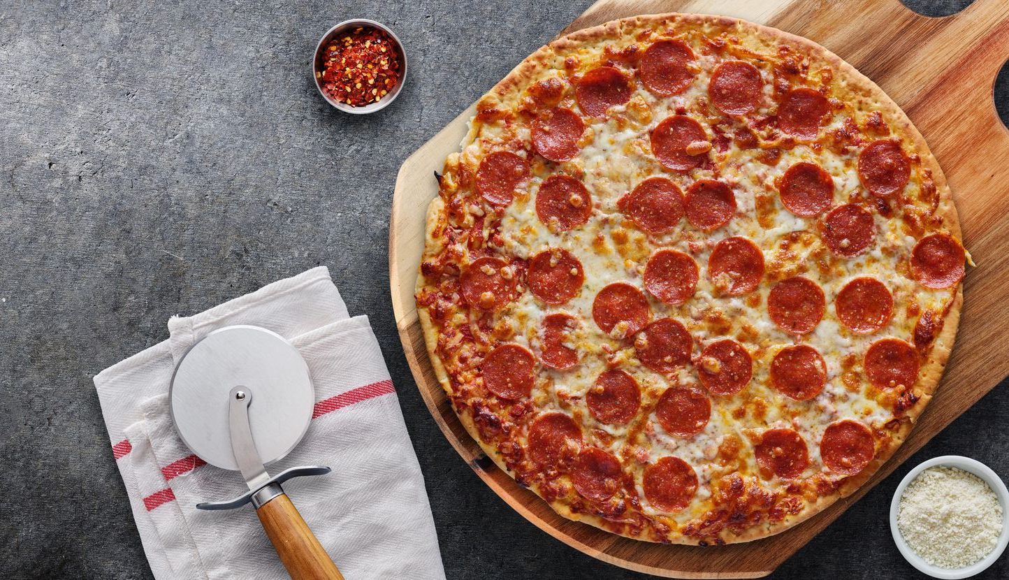 Pepperoni pizza on a wooden board with pizza cutter, red pepper flakes, and grated cheese on a gray surface