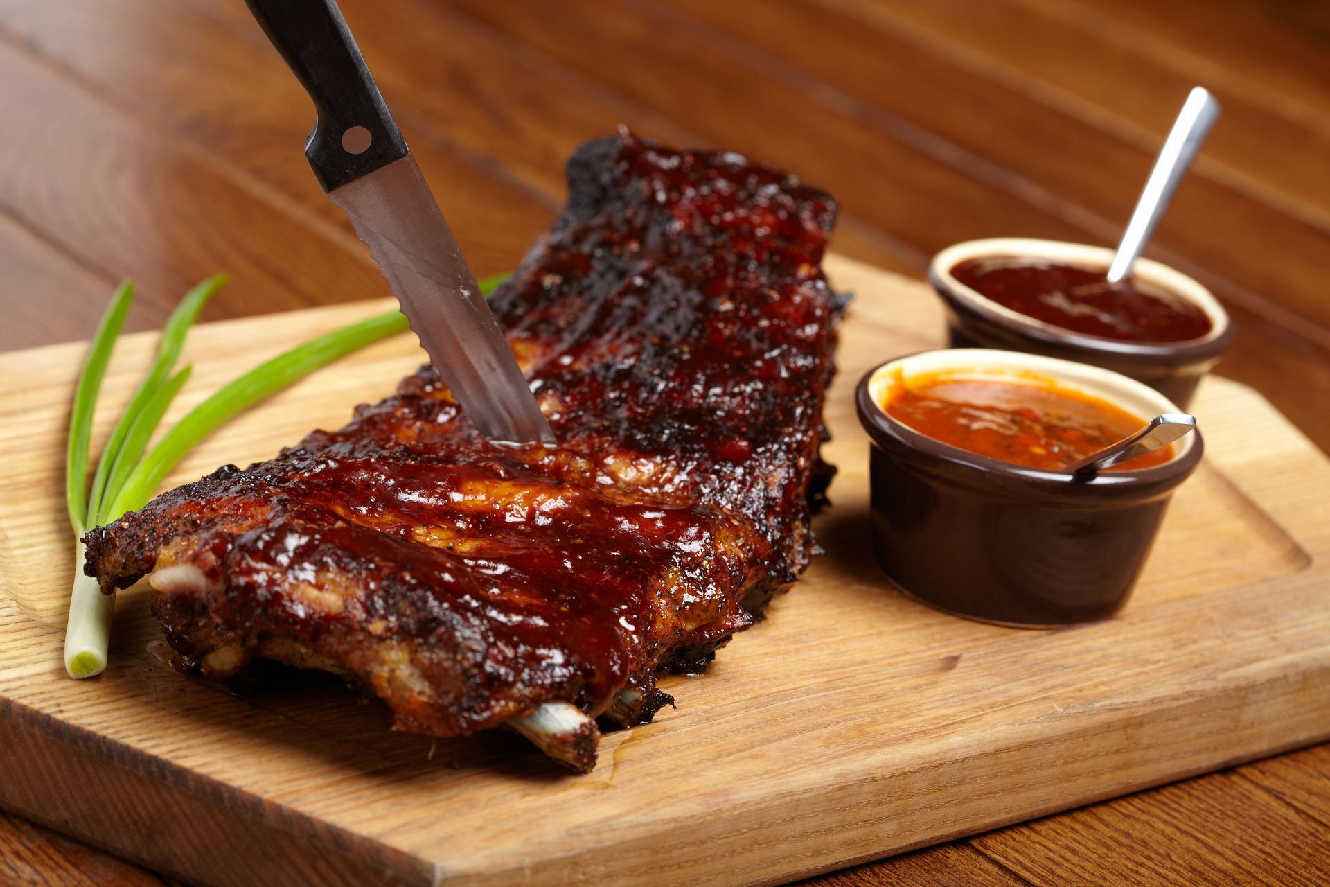 Barbecue ribs being cut on a wooden board, with sauce cups and green garnish beside them