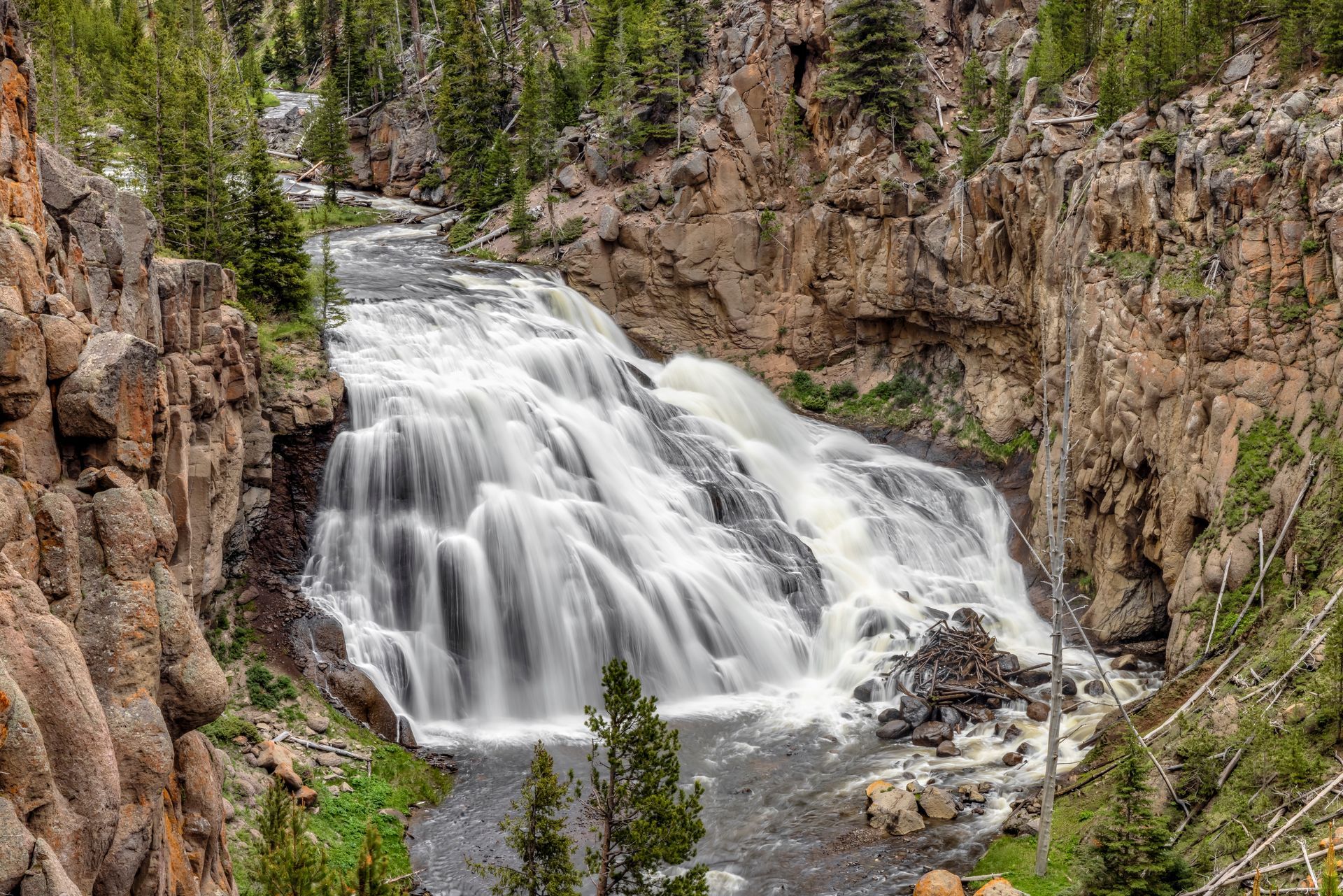Waterfall cascading over layered rock formations, surrounded by a forest in a canyon setting.
