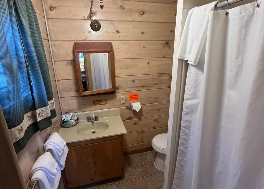 Bathroom with wood-paneled walls, a sink, toilet, shower with white curtain, and a mirror. Towels are stacked on a rack.
