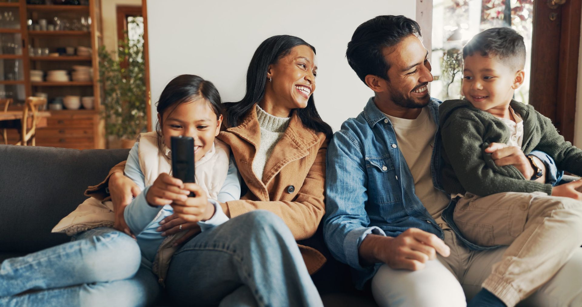 A family of four sitting on a couch in a living room, smiling as they look at a smartphone held by a child.