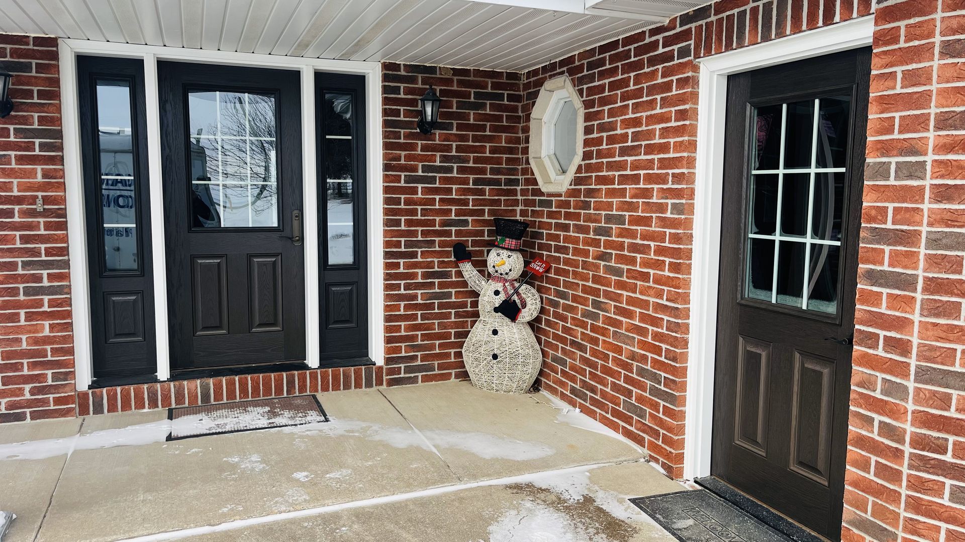 Brick building entryway with dark doors, sidelights, and a snowman decoration. Snow on the ground.
