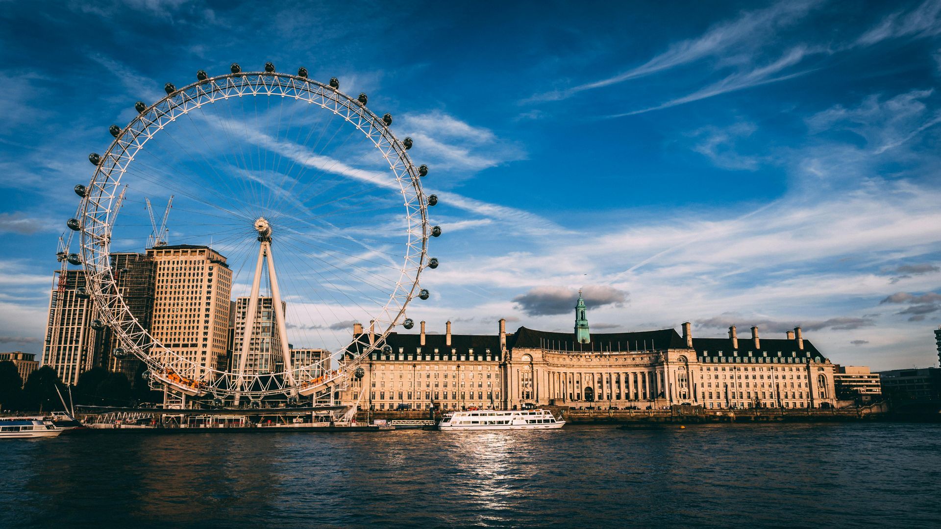 London Eye Ferris wheel and buildings along the Thames River under a blue sky.