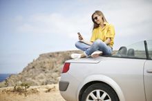 Woman sits on silver convertible car, using phone, smiles against a coastal backdrop.