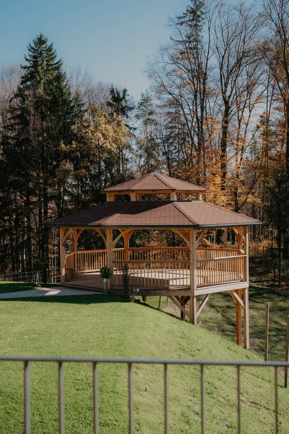 Ein hölzerner Pavillon auf einem grasbewachsenen Hügel, umgeben von Bäumen mit herbstlichem Laub.