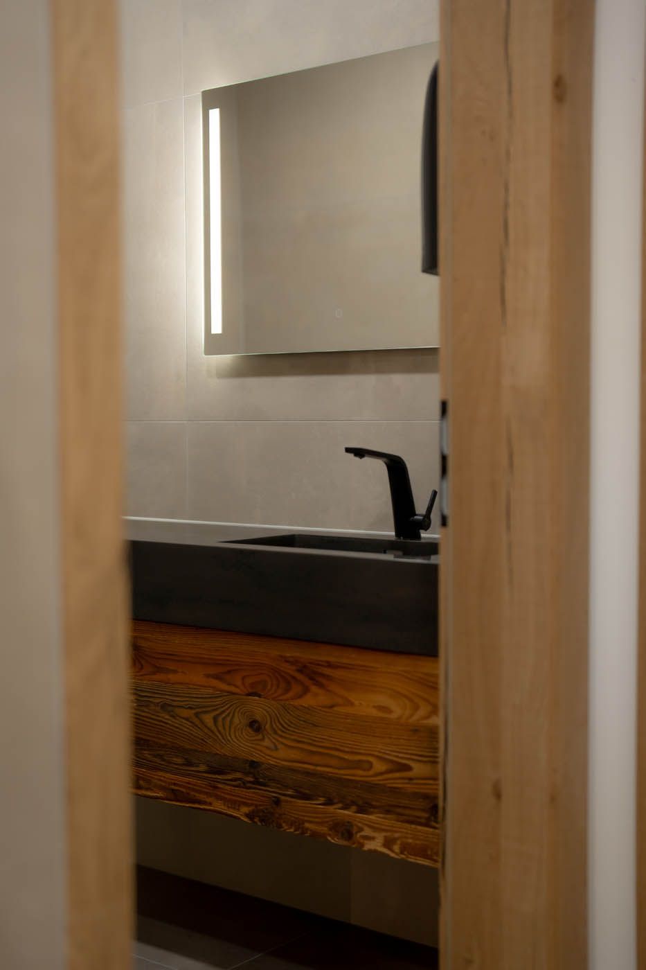 Badezimmer mit Holzschrank, dunklem Waschbecken, schwarzem Wasserhahn, beleuchtetem Spiegel, eingerahmt von Holzbalken.