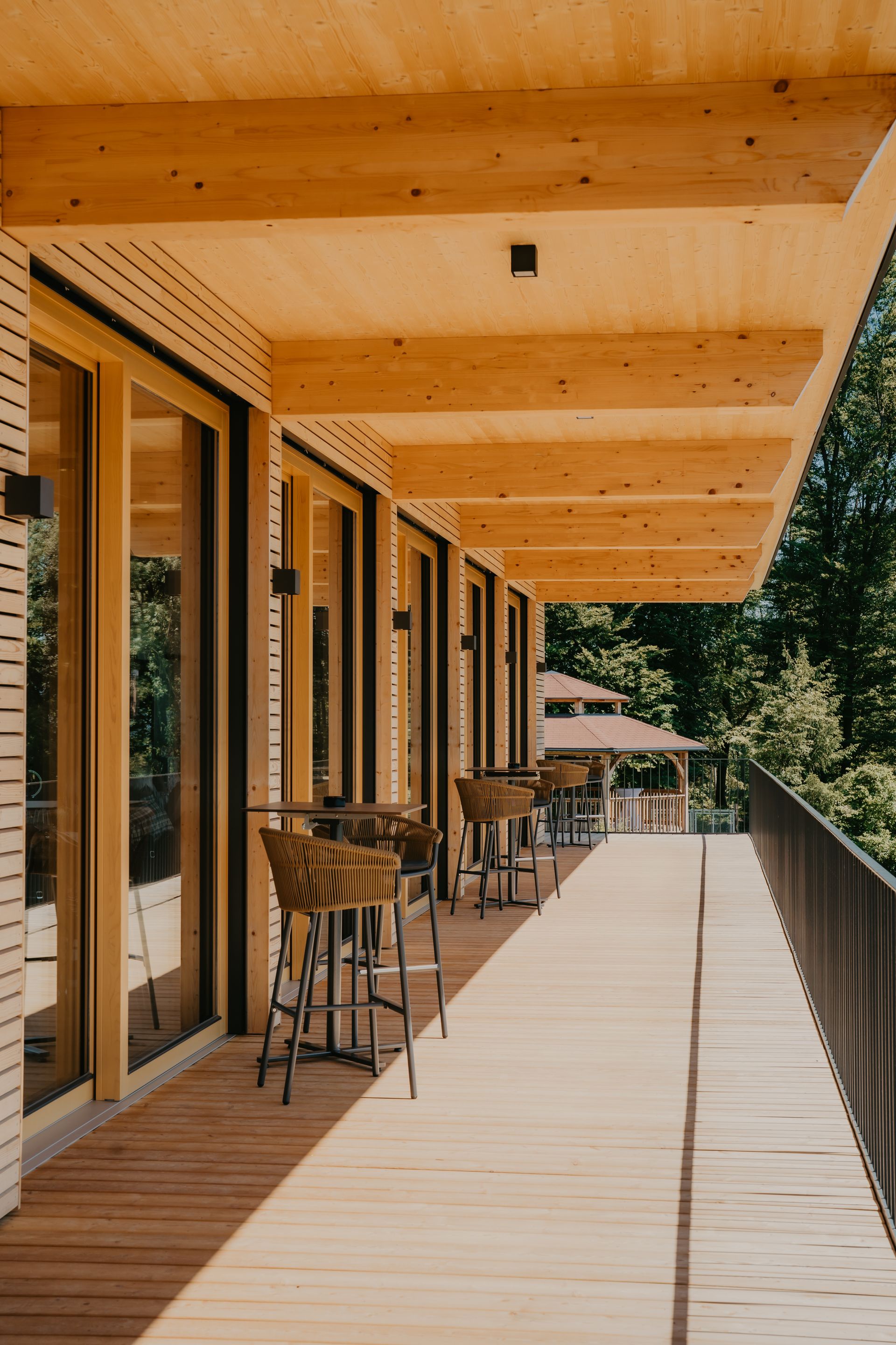 Holzterrasse mit großen Fenstern, die den Blick auf einen Pavillon inmitten eines Waldes freigeben.