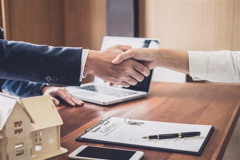 Real estate contract signing with handshake, house model, and documents on a wooden desk surface.