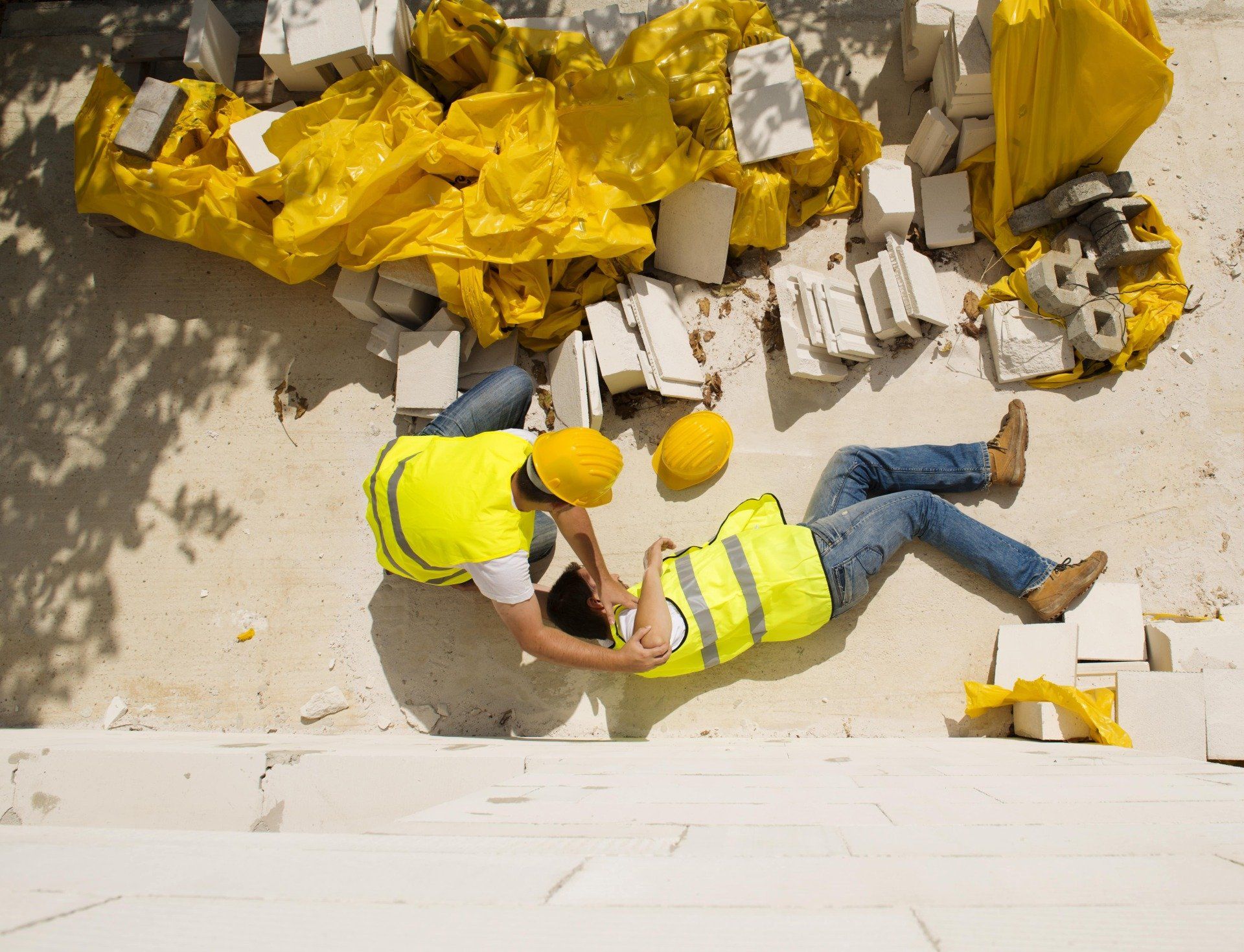 Un ouvrier du bâtiment aide un collègue blessé sur un chantier ; tous deux portent des gilets de sécurité et des casques de chantier.