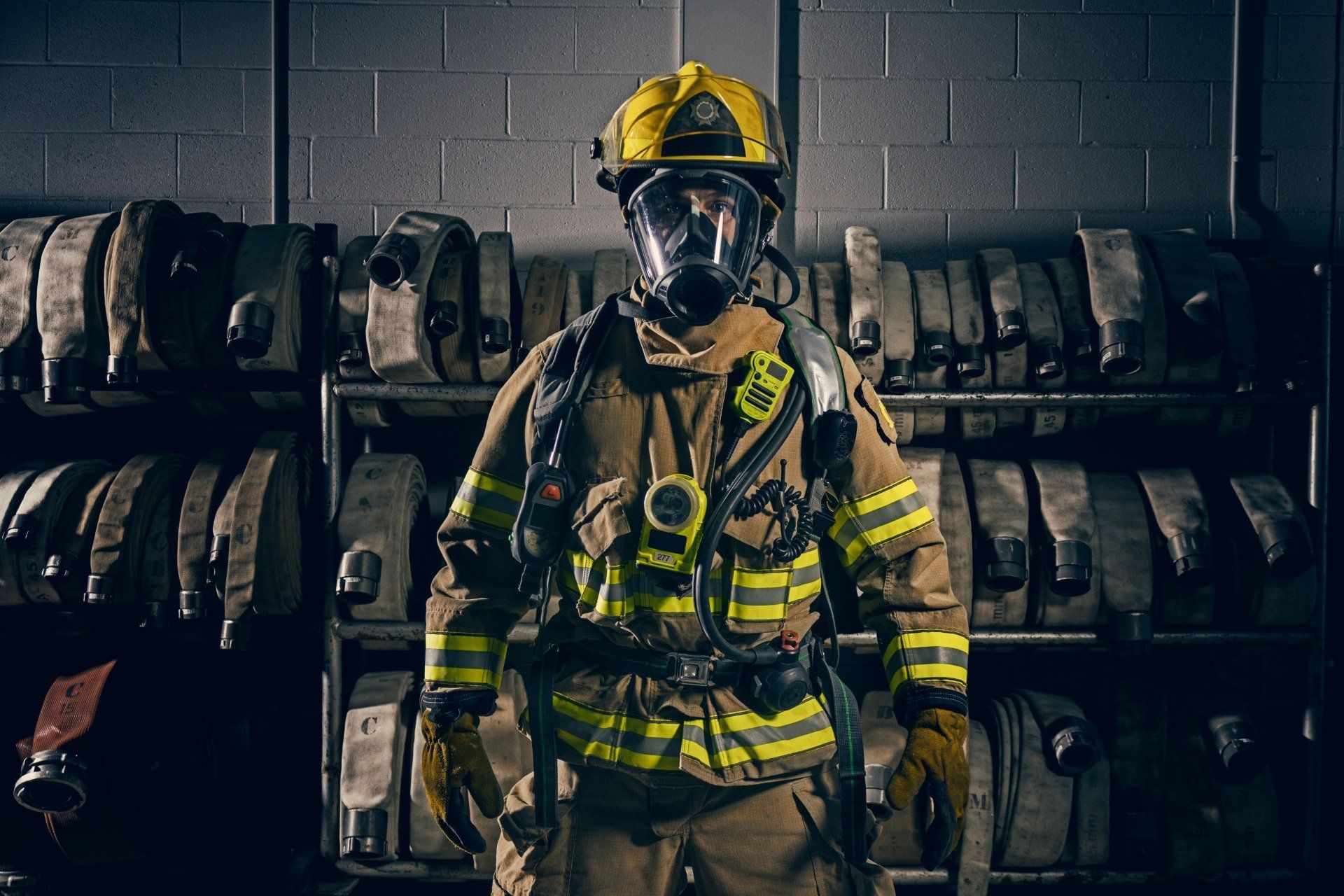 Firefighter in yellow helmet and gear, wearing a respirator, standing in front of hose racks in a dim room.