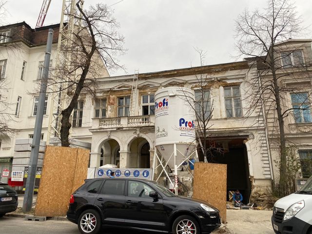 Gebäude im Umbau; weißer Silo, schwarzes Auto und Sperrholzbarrieren davor.