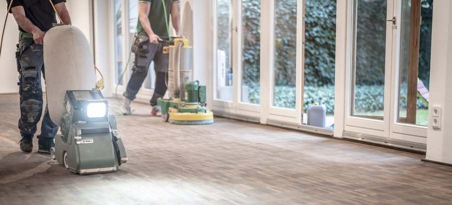 Deux ouvriers polissent un parquet dans une pièce lumineuse dotée de grandes fenêtres et d'une polisseuse.