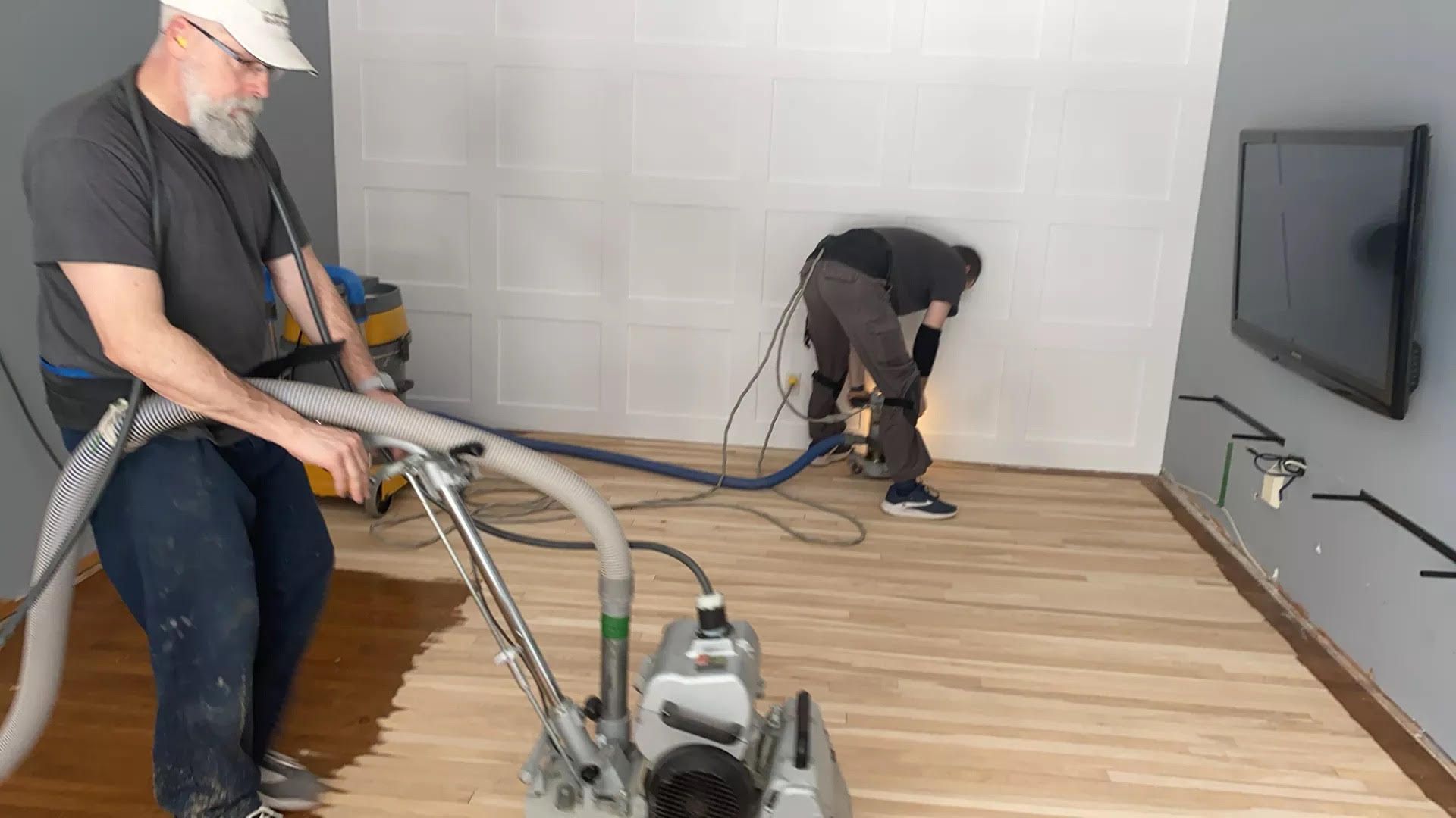 Deux ouvriers utilisent des ponceuses à parquet sur un plancher en bois dans une pièce où se trouve une télévision fixée au mur.