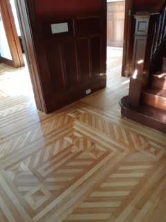 Couloir avec parquet à motifs géométriques et lambris en bois foncé, éclairé par la lumière du jour provenant d'une porte