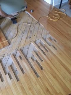 Une personne, allongée sur un plancher en bois, utilise un pistolet thermique pour décaper la peinture d'une section de planches.