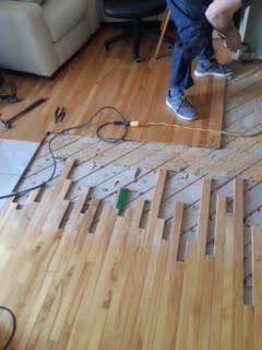 Plancher en bois franc endommagé, avec des lames et des outils manquants, tandis qu'une personne est agenouillée à proximité dans une pièce