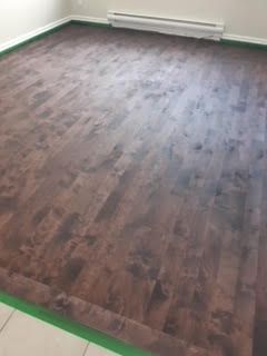 Pièce vide avec un sol imitation bois foncé et des plinthes blanches, partiellement recouvertes de ruban adhésif de peintre vert.