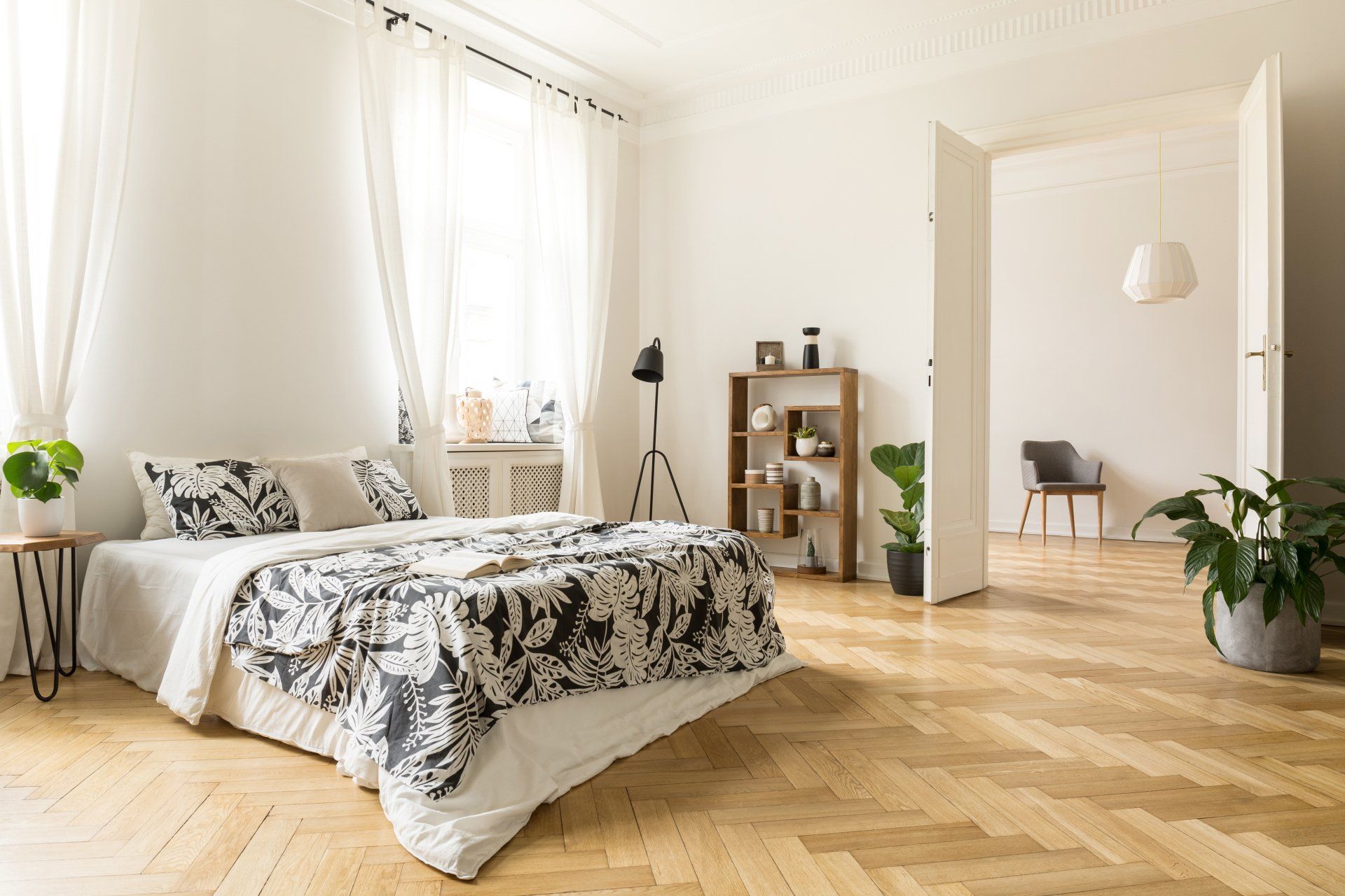 Chambre lumineuse avec un lit, des voilages, un parquet et des plantes en pot près d'une porte ouverte.