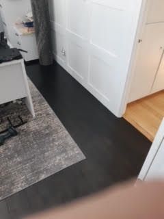 Couloir avec sol foncé, tapis gris, murs blancs et porte ouverte donnant sur une pièce au parquet.