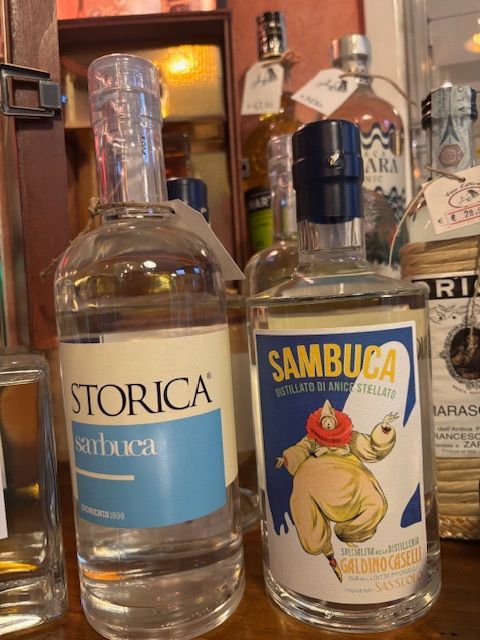 Su uno scaffale ci sono due bottiglie di liquore: una bottiglia di Torica trasparente e una di Sambuca con l'etichetta blu Vino Estremo Enoteca Soliera Apuana e La Spezia