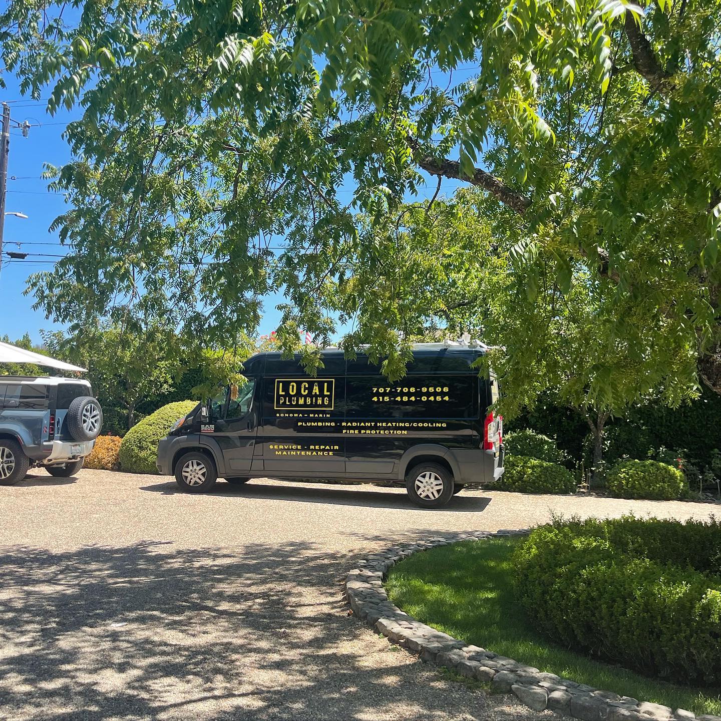 Dark gray Local Plumbing van parked on a driveway, trees overhead, another vehicle in view.