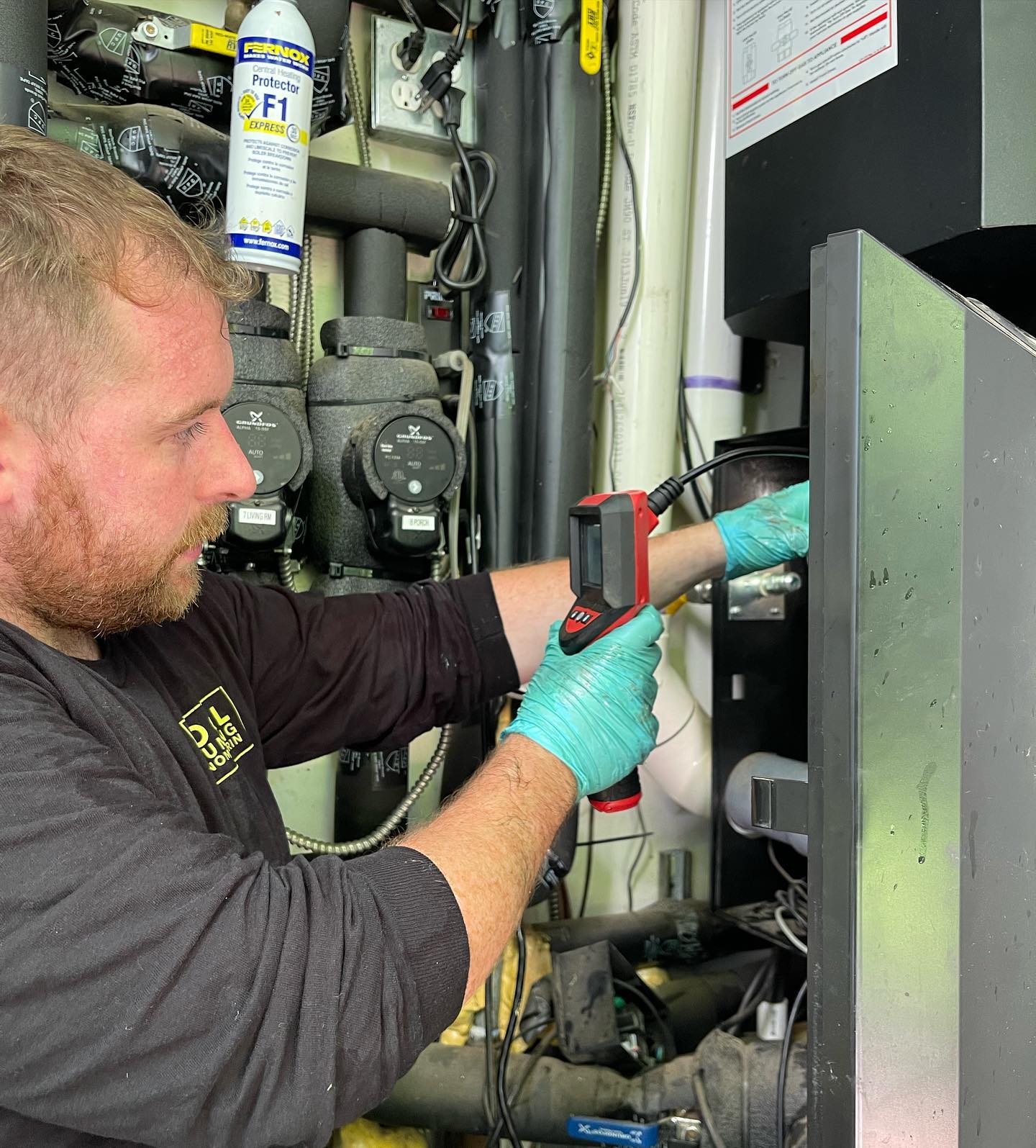 A person in a black shirt and gloves uses a handheld thermal camera to inspect a mechanical system.
