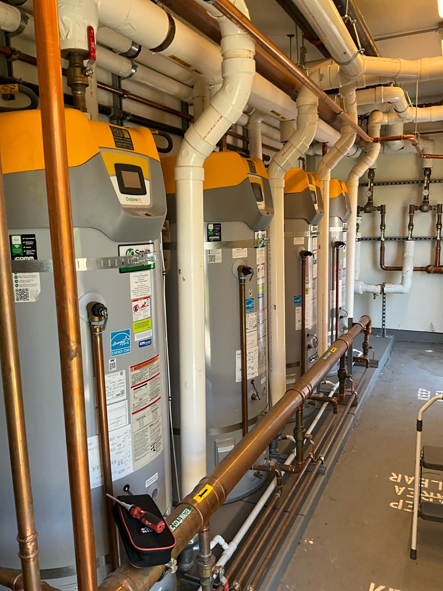 Water heaters lined up in a utility room with pipes and equipment.