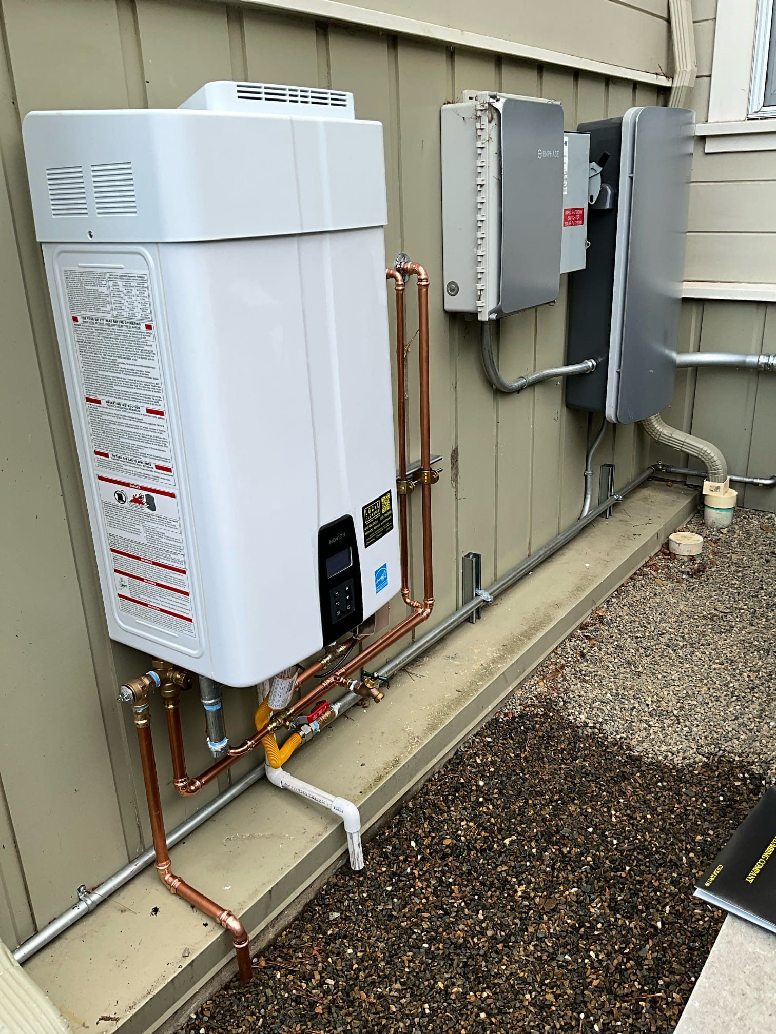 White tankless water heater, copper pipes, and electrical boxes mounted on a beige exterior wall.