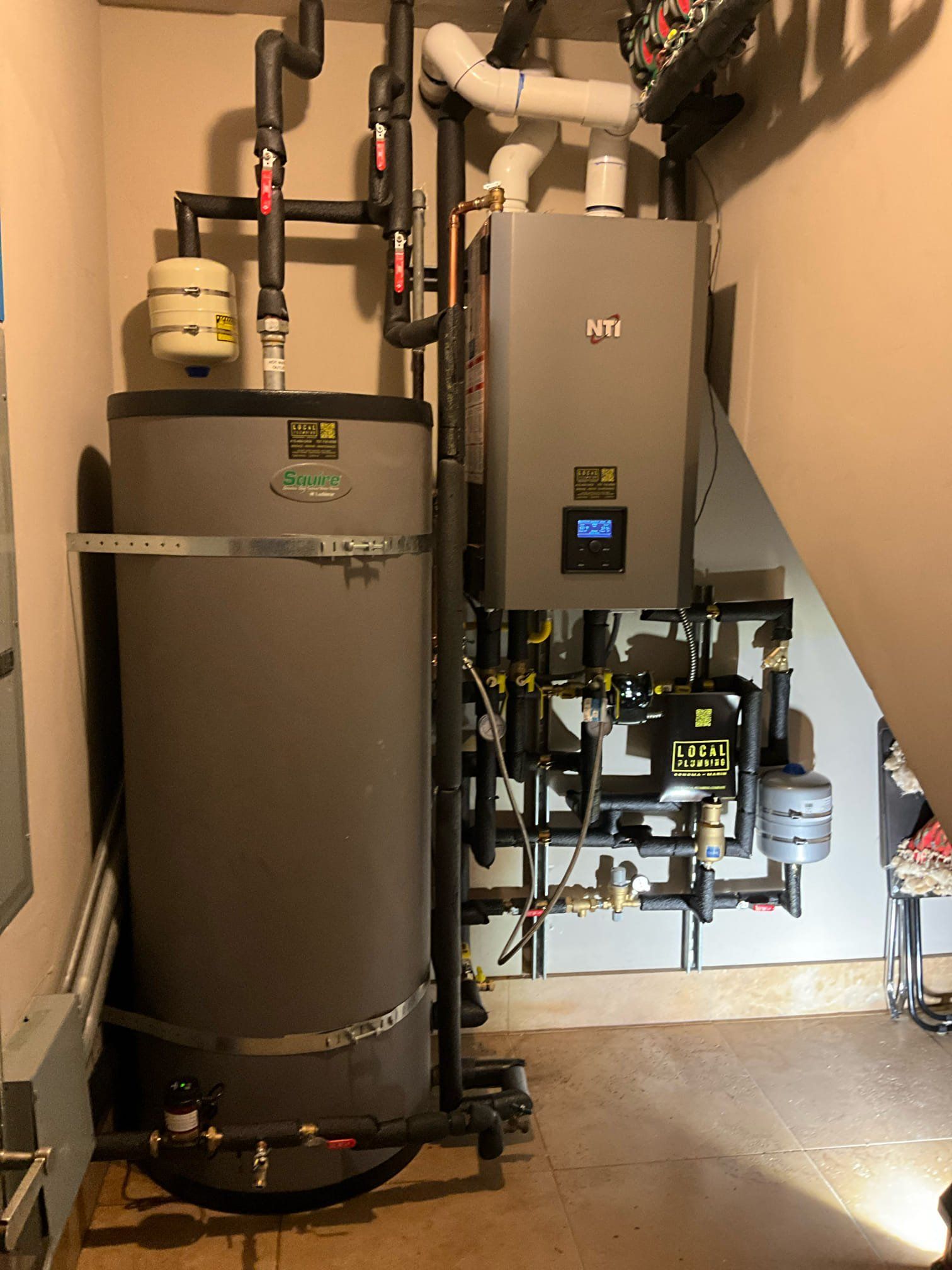 A residential water heating system with a tall gray tank, and a smaller wall-mounted unit. Pipes and controls are visible.