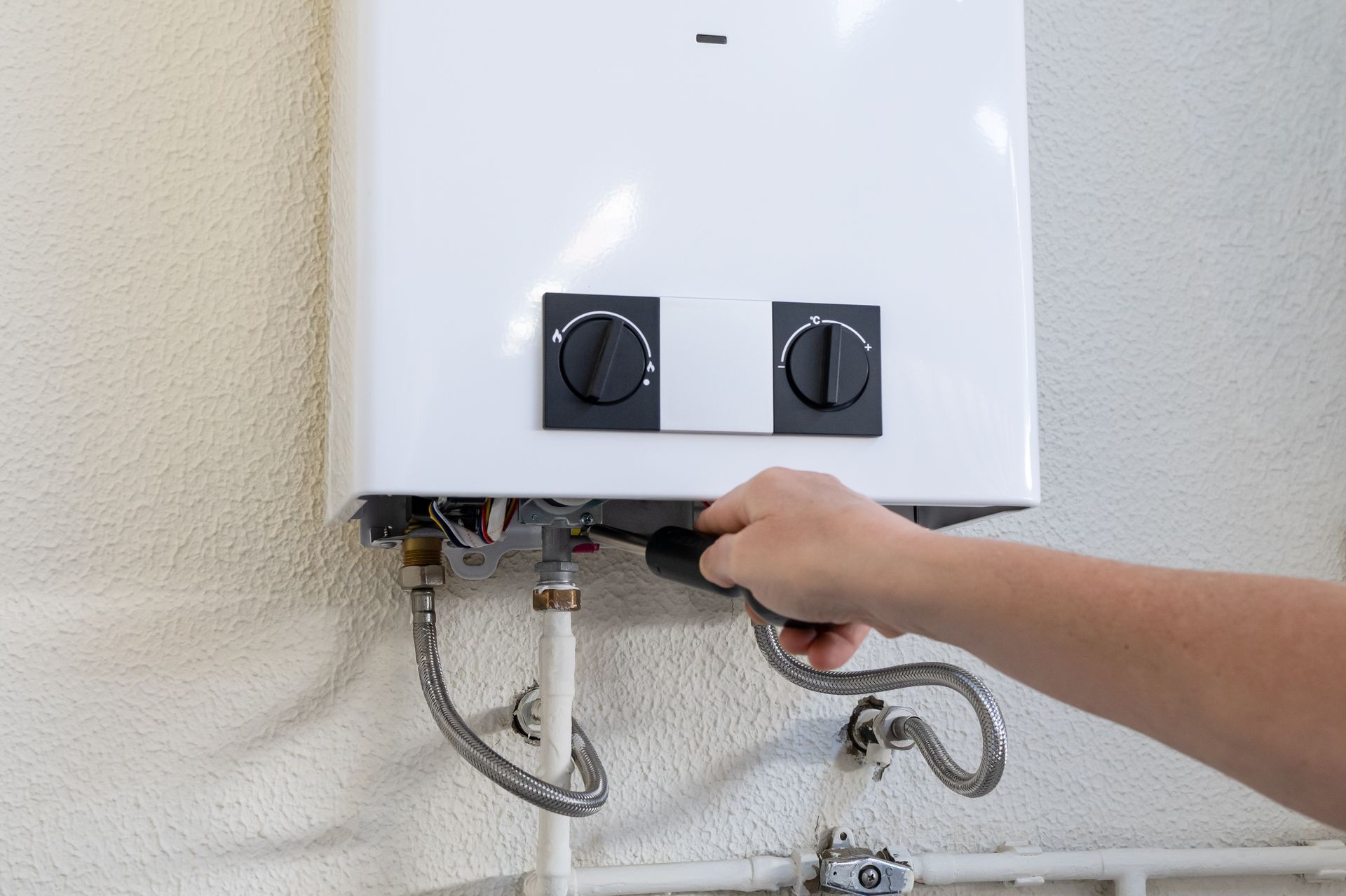 A plumber in a blue uniform and gray gloves uses an orange pipe wrench to tighten a chrome sink Hand connecting a flexible hose to a wall-mounted tankless gas water heater.