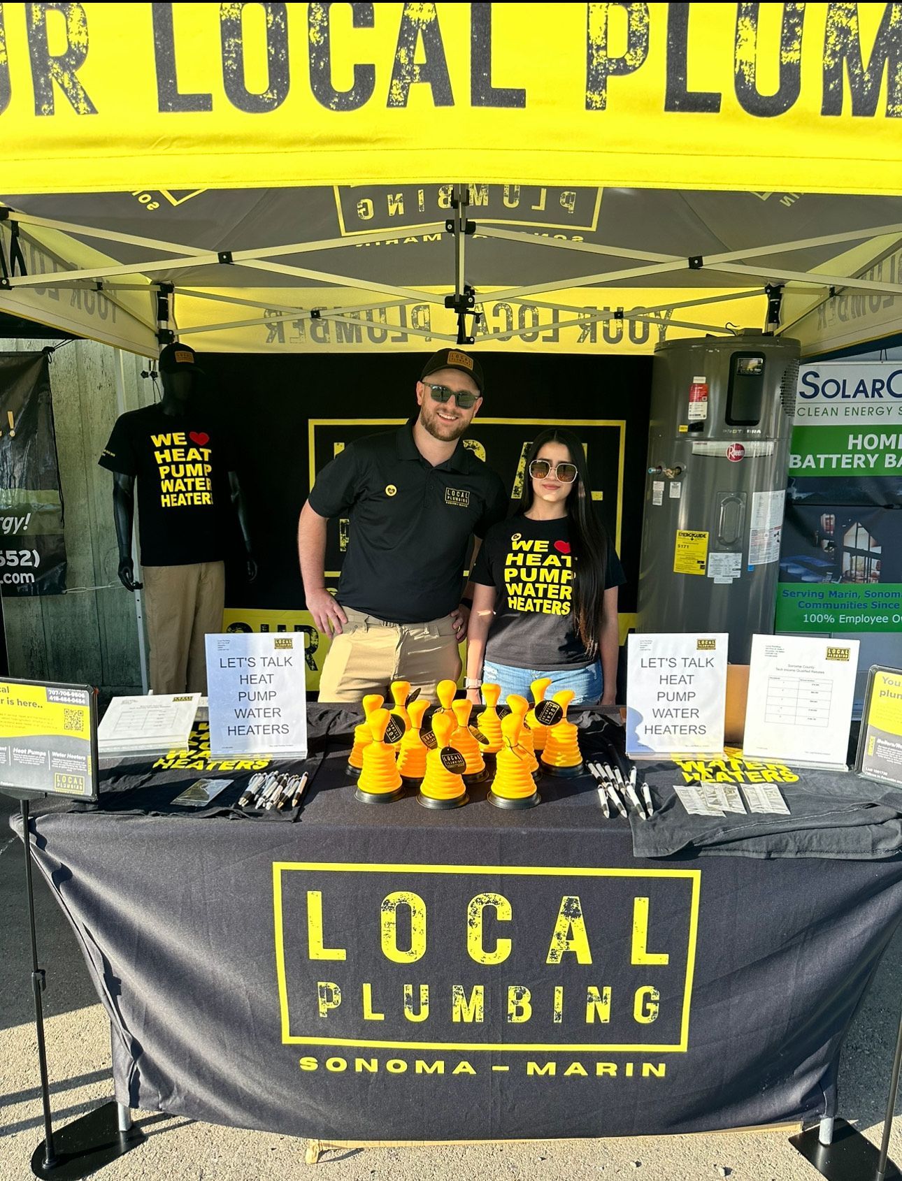 Local plumbing booth at an event; man and woman standing behind a table with promotional items, banner reads