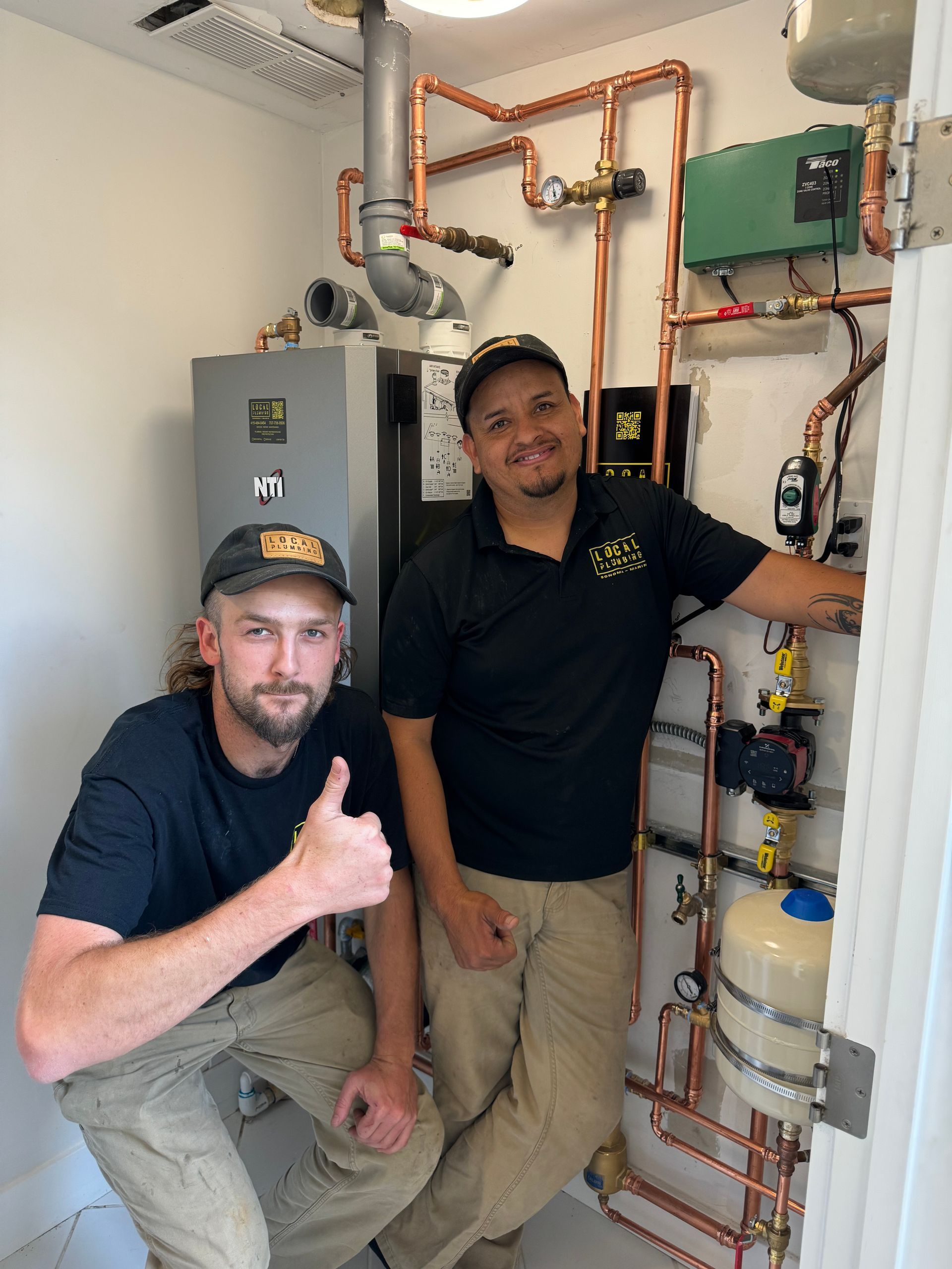 Two workers by heating system. One gives thumbs up. Copper pipes, gray tank, white door frame.