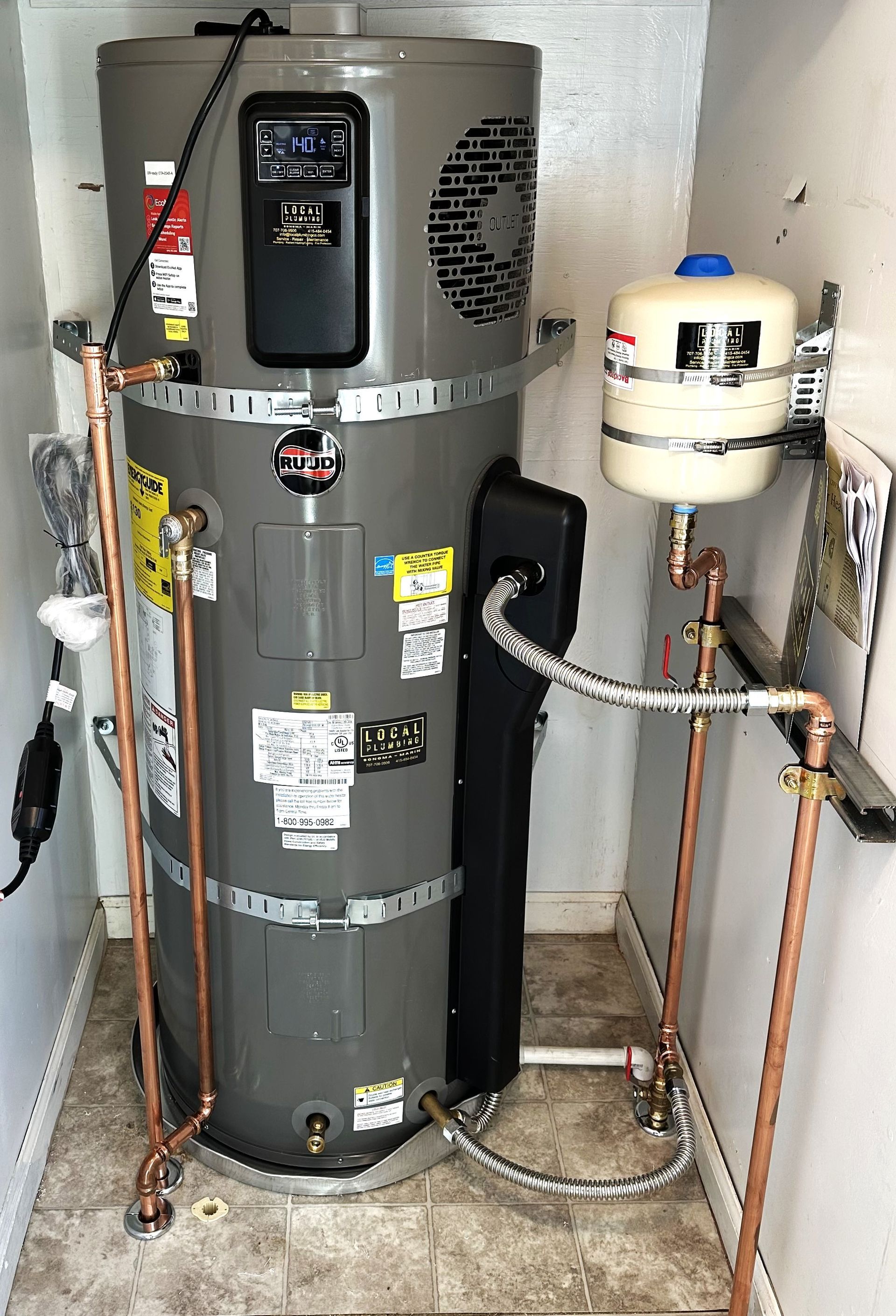 A water heater and attached expansion tank in an enclosed space with copper pipes and various labels.