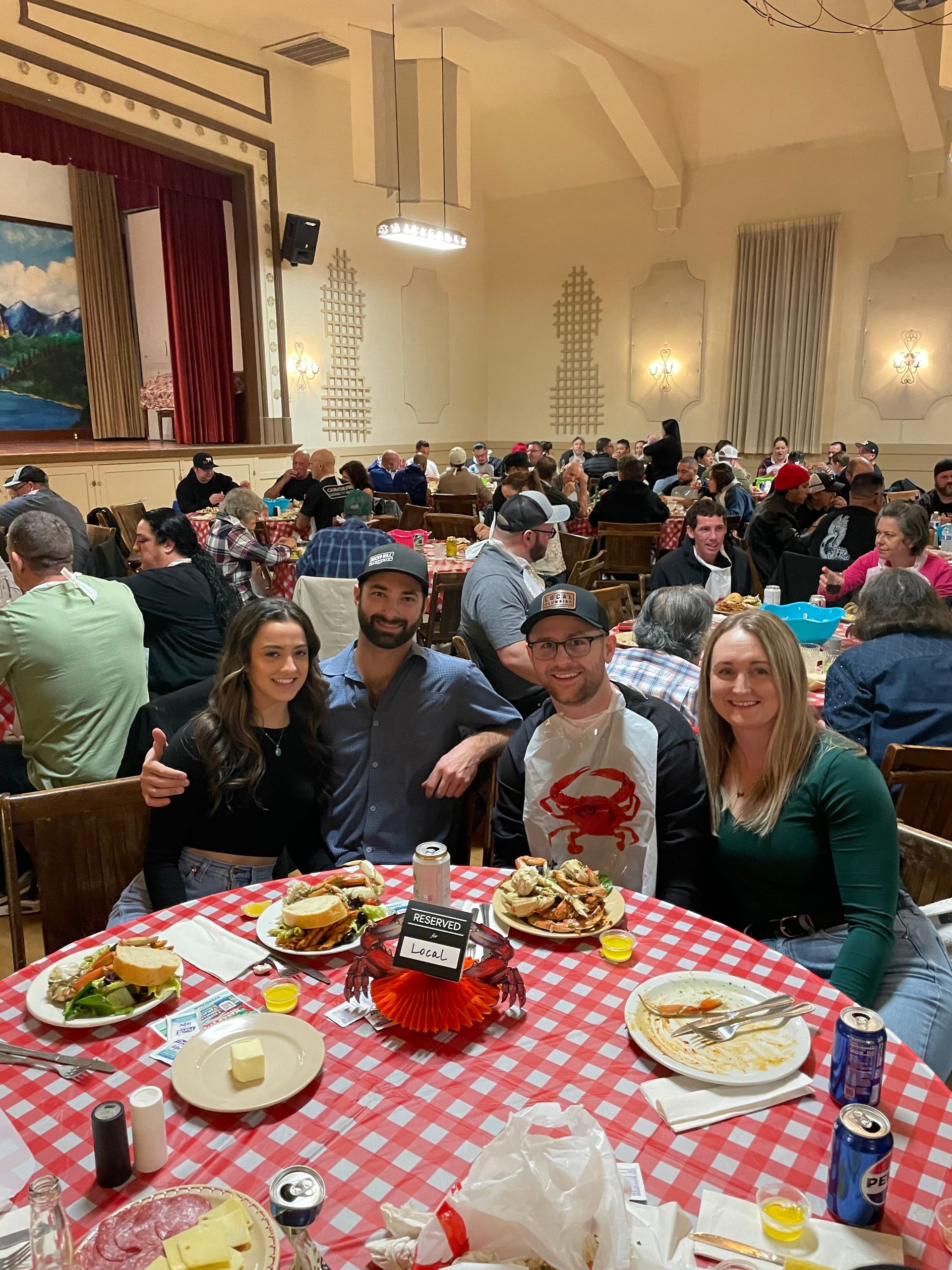 Four people seated at a table covered in a red-and-white checkered tablecloth, enjoying food in a crowded hall.