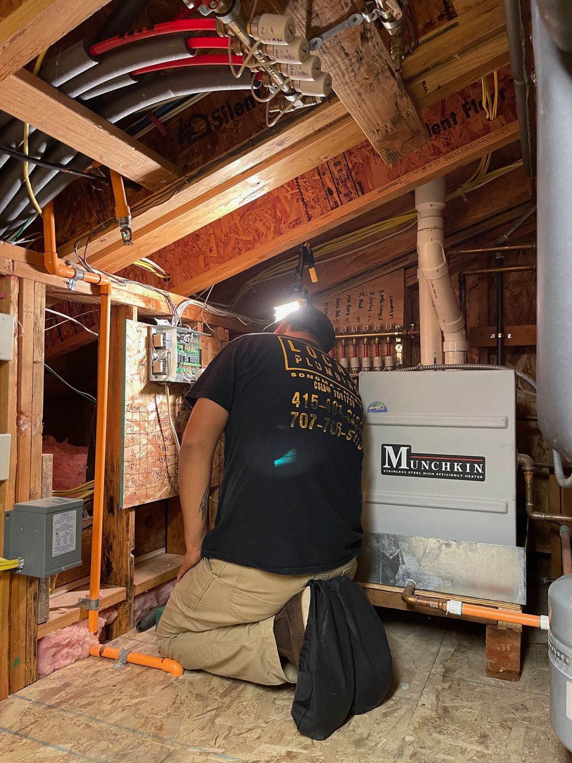Person in a black shirt working on electrical components inside an attic. Plumbing and HVAC equipment visible.