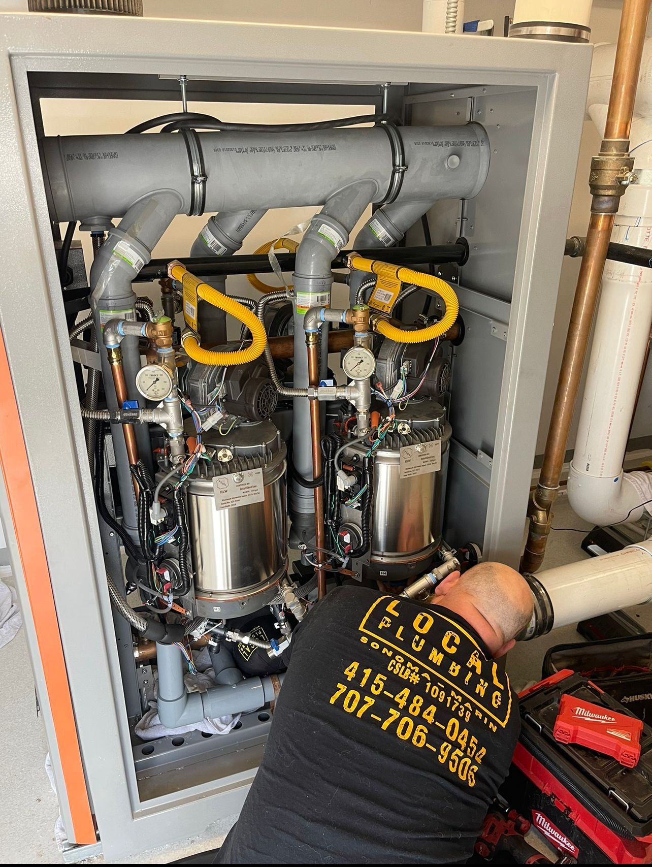 Plumber working on a complex water heater system inside a cabinet. Yellow piping, gray cabinet, and tools visible.