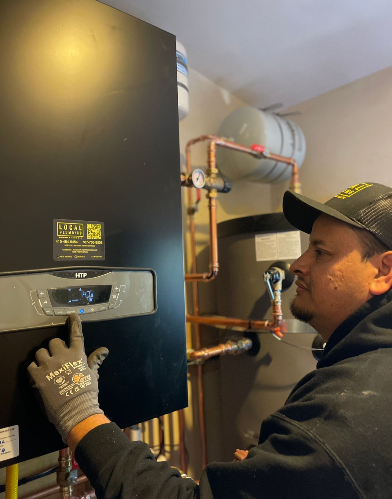 Person in a cap and work gloves adjusting a heating system's control panel; copper pipes and tank in the background.