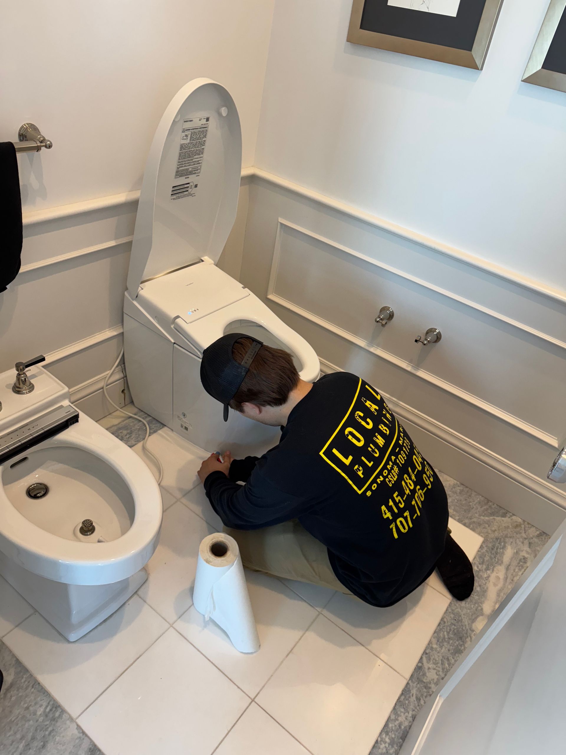 Plumber working on a toilet and bidet in a bathroom. He is kneeling, working on the toilet, with a roll of toilet paper.
