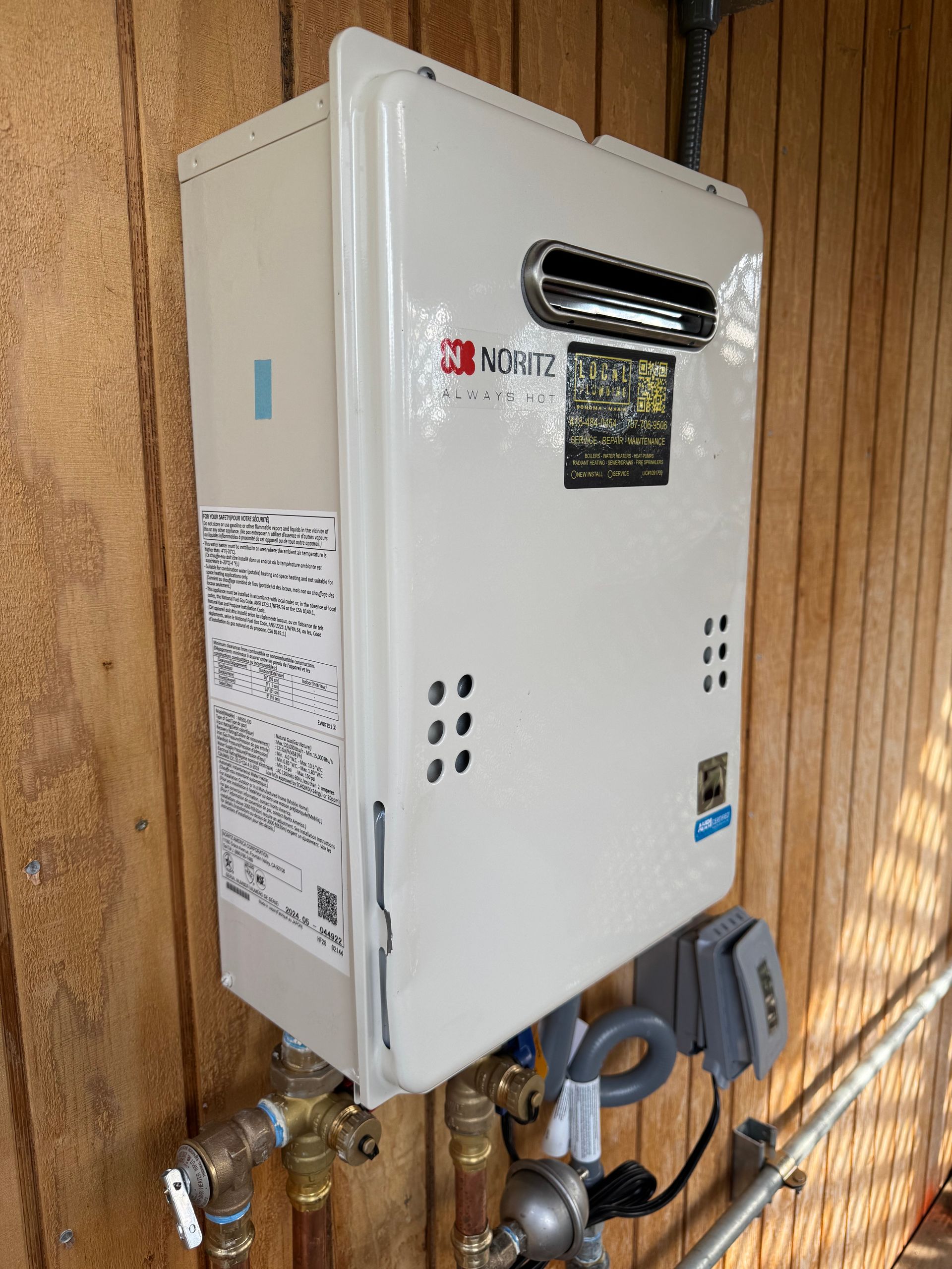 White Noritz tankless water heater mounted on a wood-paneled wall, with copper pipes and gray control box.