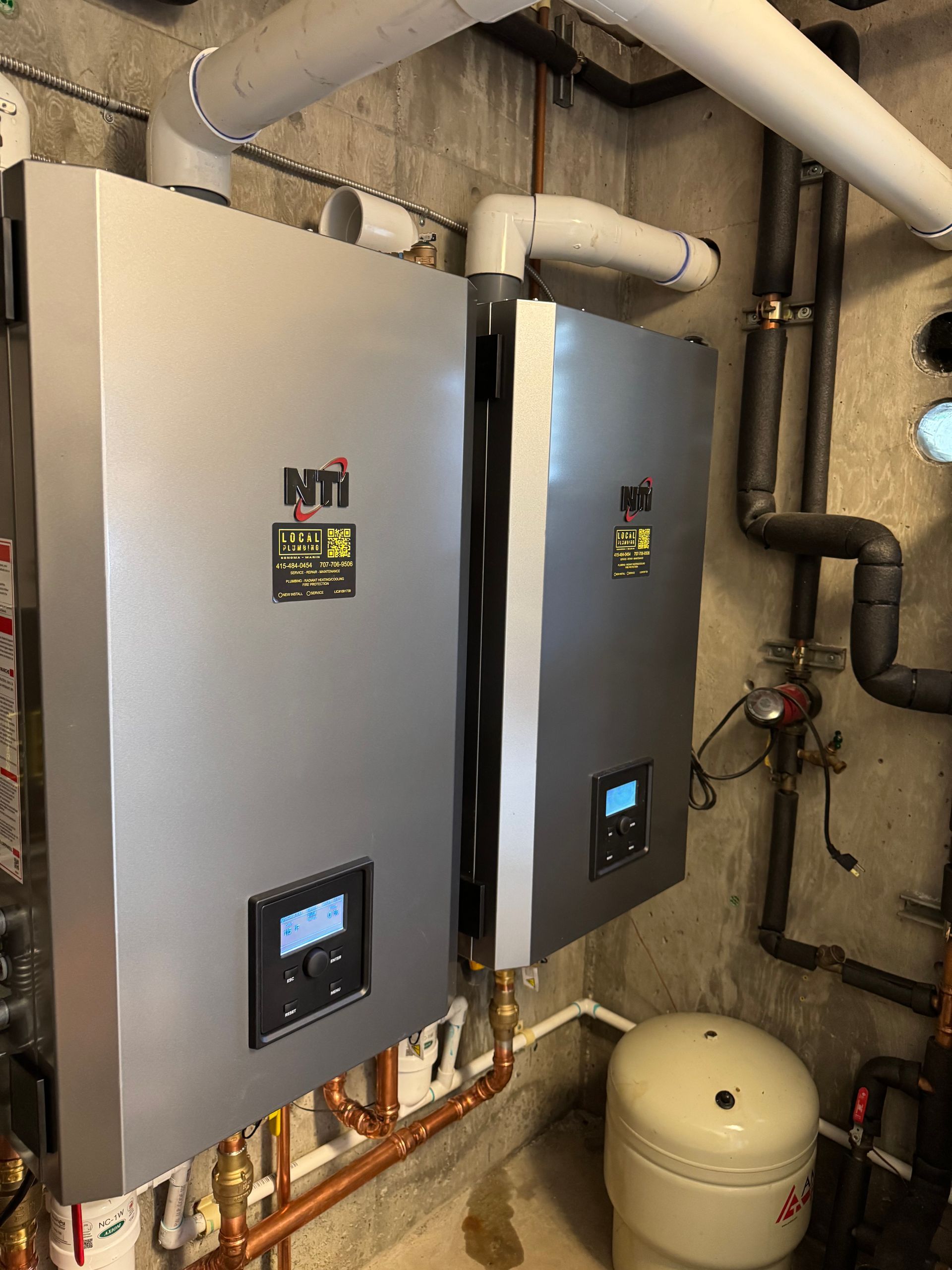 Two silver NTI boilers mounted on a concrete wall with copper piping and white ventilation pipes.