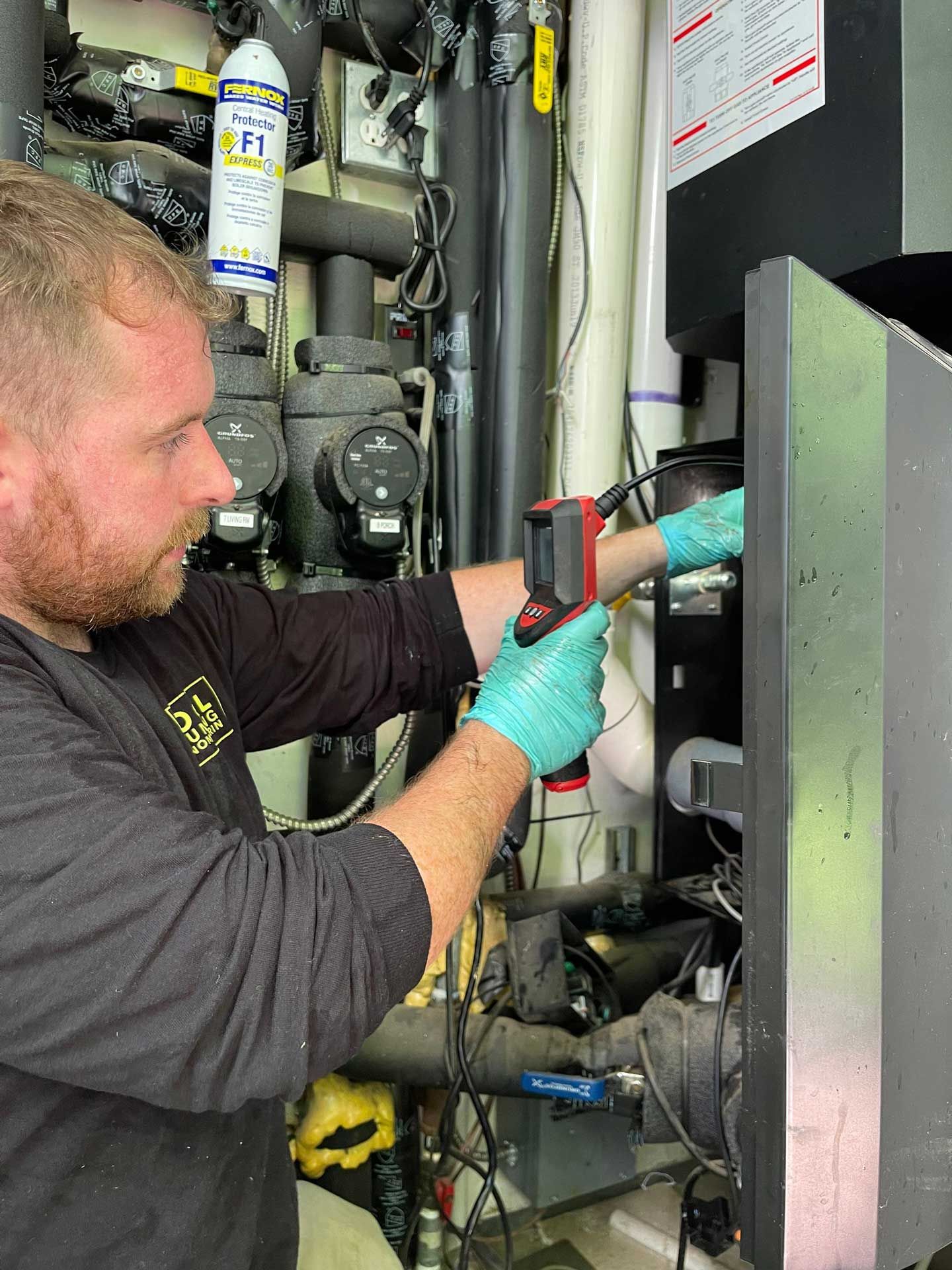A person in gloves uses a temperature gun on machinery in an indoor utility space.