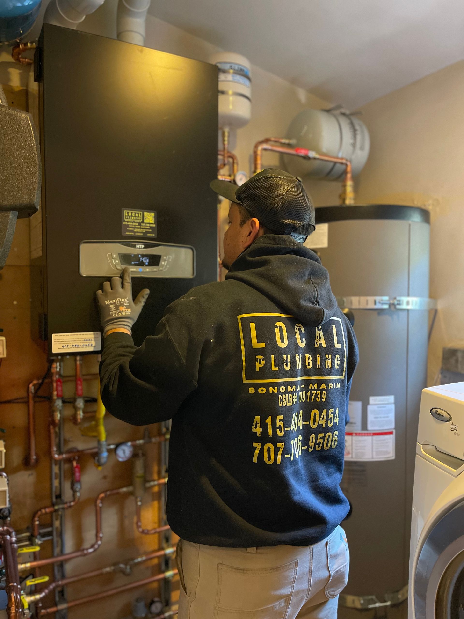 Plumber in a black hoodie installing a water heater in a utility room. Copper pipes and a washing machine are visible.