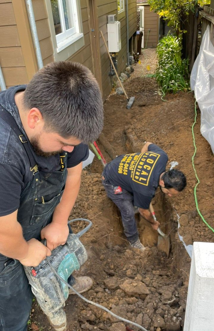 Two people digging a trench outdoors. One uses a jackhammer, the other a shovel. Brown soil.